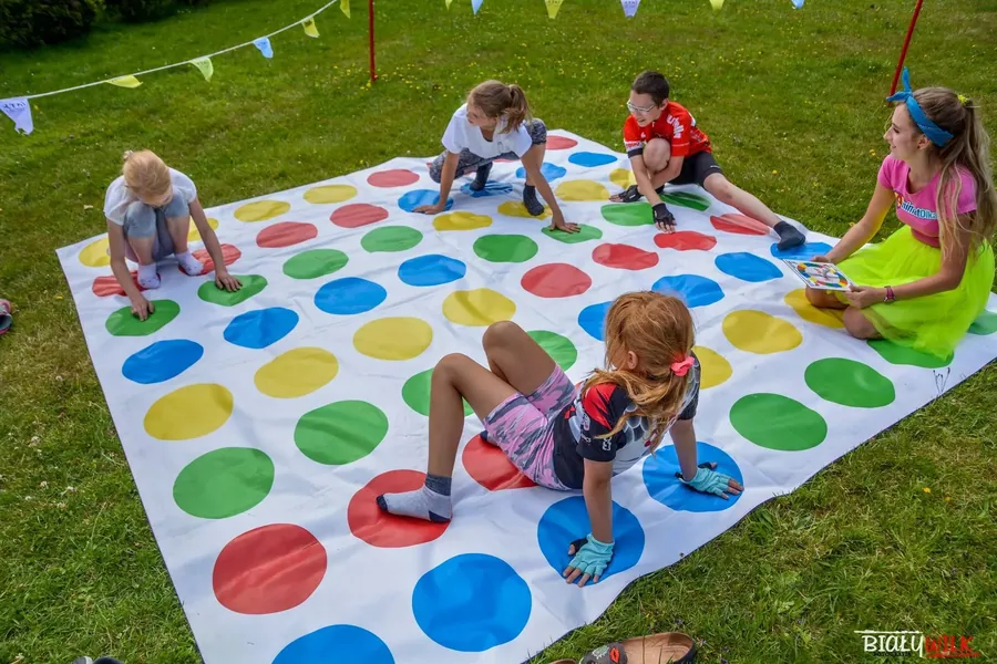 Wielki Twister rozłożony na trawie, dzieci i dorośli grają razem